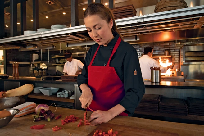 Restaurant Uniform Rentals - Chef in black chef coat and red apron dicing vegetables